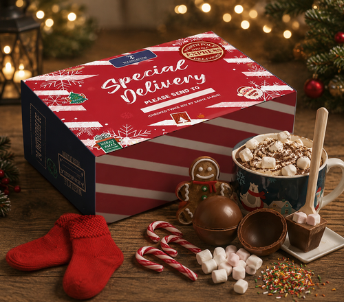 Tilz Collection Christmas Eve Box for Adults with contents displayed including festive mug filled with hot chocolate and marshmallows, gingerbread man biscuit, chocolate bombs, candy cane, red Christmas socks, sprinkles and wooden stirrer on festive wooden table